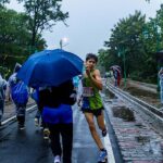 A marathon runner in a green vest with bib 5156 smiles while running on a wet road, passing spectators with umbrellas during the Pachmarhi Monsoon Marathon.