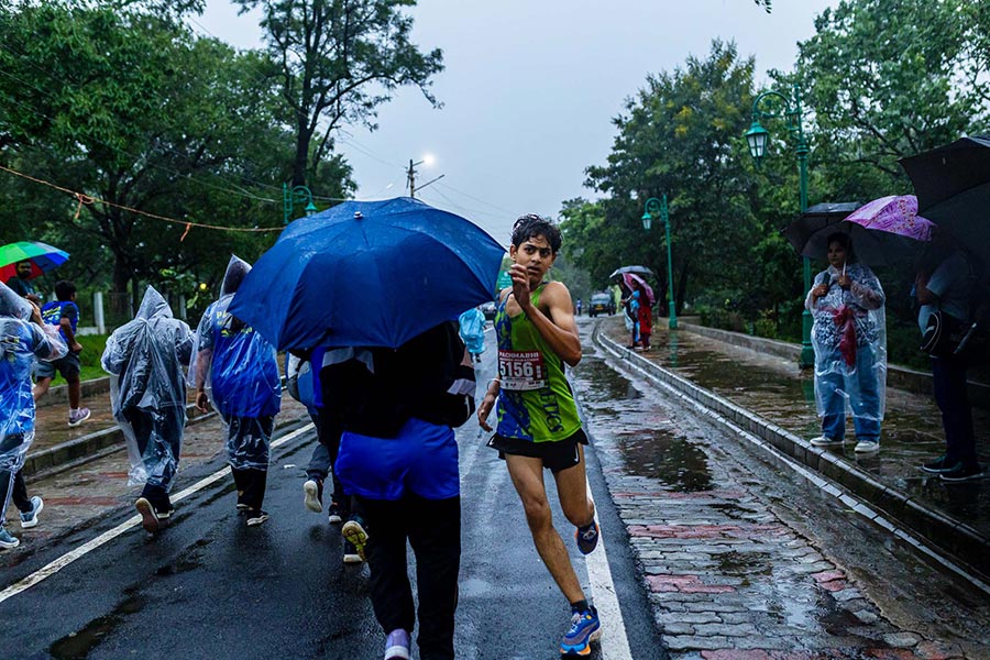 A marathon runner in a green vest with bib 5156 smiles while running on a wet road, passing spectators with umbrellas during the Pachmarhi Monsoon Marathon.