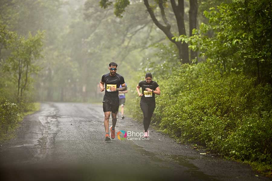 Two marathon runners, a man and a woman, run on a wet, misty road surrounded by lush green trees during the Pachmarhi Monsoon Marathon in the Satpura Hills.
