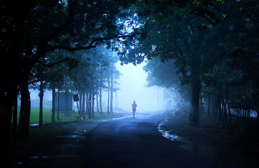 A lone marathon runner in a red shirt runs into a dense, atmospheric mist on a dark, wet road surrounded by trees, captured during the Pachmarhi Monsoon Marathon.